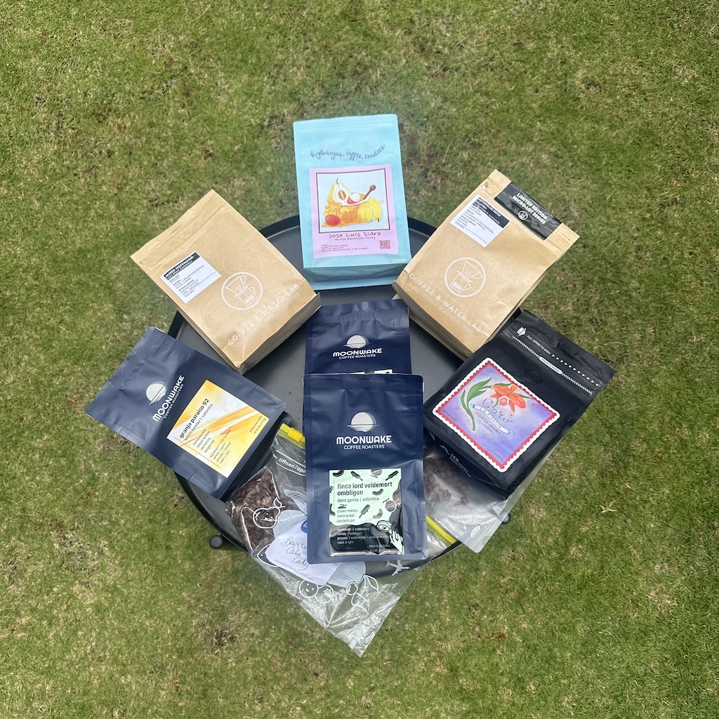 An image showing about 9 bags of coffee from CoRo, Moonwake, Coffee and Water Lan, and Hydrangea roasters on a black Ikea stand with grass at the background. All beans are sourced from the americas.